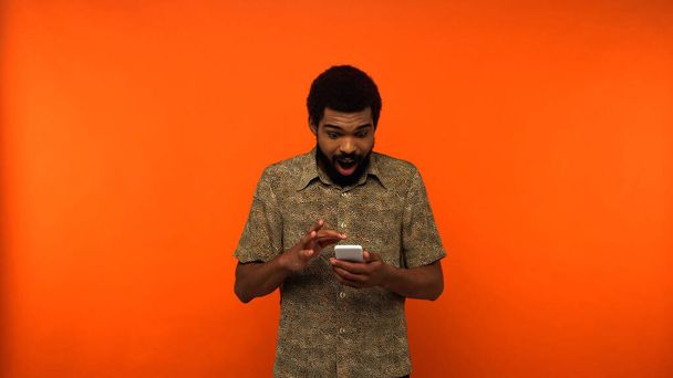 hämmästynyt afrikkalainen amerikkalainen mies käyttää älypuhelinta oranssilla taustalla  - Valokuva, kuva