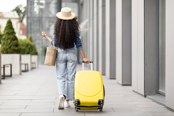 Tourism concept. Unrecognizable brunette long-haired woman in casual outfit having journey abroad, walking by modern airport with yellow luggage and straw bag, panorama with copy space, full length - Photo, Image