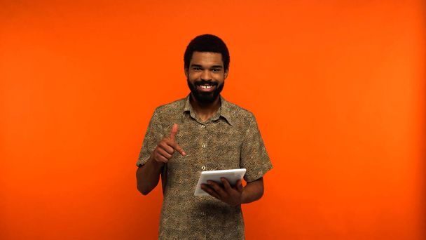 boldog afro-amerikai férfi szakállas ujjal mutat a digitális tabletta narancs alapon  - Fotó, kép