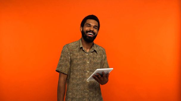 szczęśliwy Afrykański Amerykanin z brodą trzymający cyfrową tabletkę i śmiejący się na pomarańczowym tle  - Zdjęcie, obraz