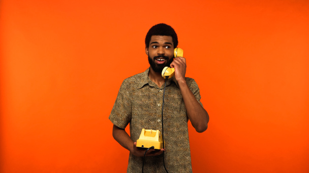 happy african american man with beard talking on yellow retro telephone on orange background  - 写真・画像