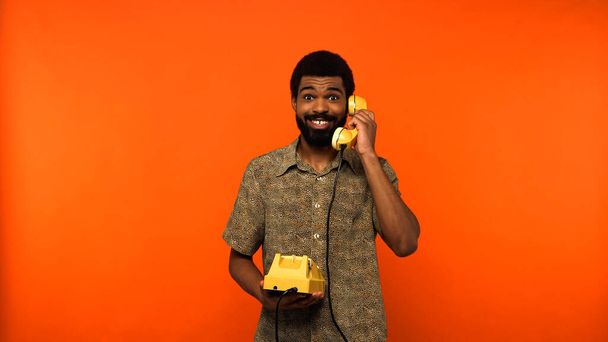 usmívající se africký Američan s vousy mluví na retro telefon na oranžovém pozadí  - Fotografie, Obrázek