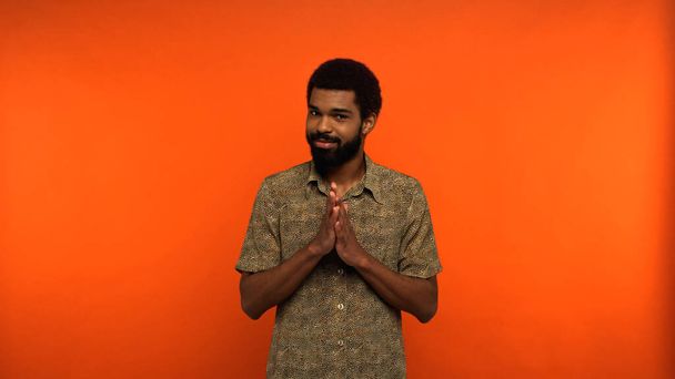 sly african american man with beard looking at camera while gesturing on orange background - Foto, Bild