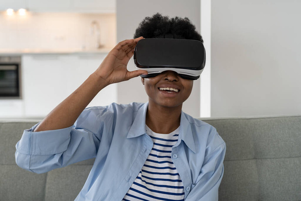 若いアフリカ系アメリカ人女性を3Dメガネで笑顔で仮想現実機能リラックスソファの上に座っています。VRヘッドセットを持つカジュアルブラックガールは、ビデオゲームや現代のガジェットへの中毒に苦しんでいます - 写真・画像