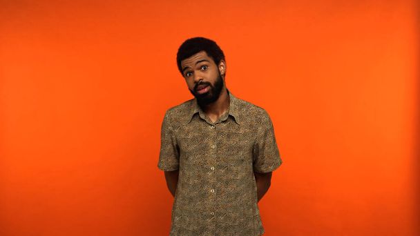 homme afro-américain perplexe avec barbe regardant la caméra sur fond orange - Photo, image