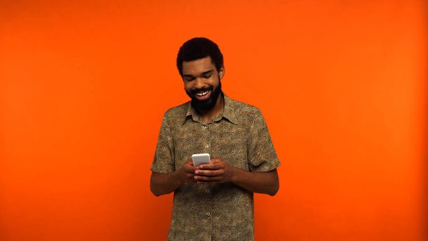 positive and bearded african american man using smartphone on orange background  - Photo, Image