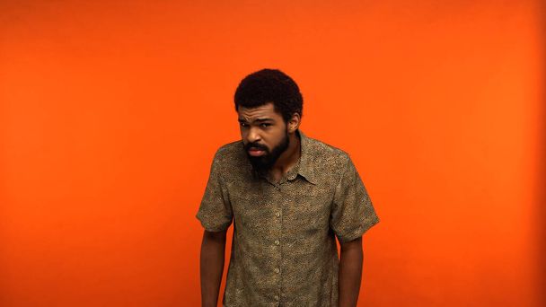 hombre afroamericano incierto en camisa mirando la cámara mientras está de pie sobre fondo naranja  - Foto, imagen