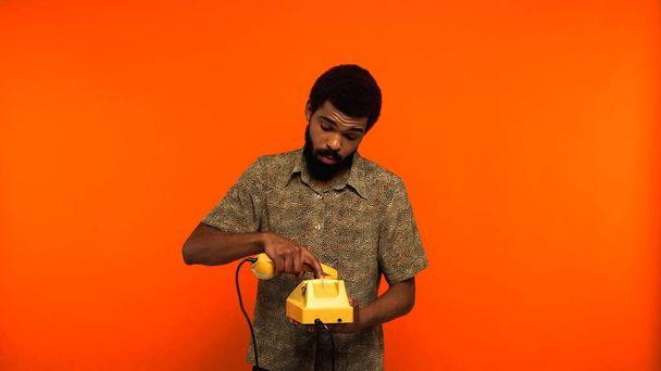 オレンジの背景に黄色のレトロな電話で髭を生やした若いアフリカ系アメリカ人男性  - 写真・画像