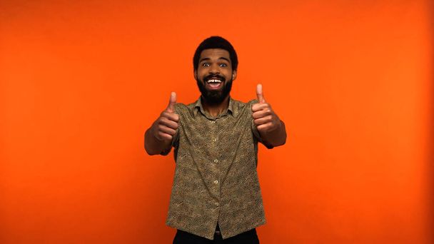 pozytywny Afroamerykanin człowiek z otwartymi ustami pokazując kciuki w górę na pomarańczowym tle  - Zdjęcie, obraz