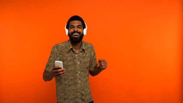 bearded african american man in wireless headphones holding smartphone on orange background  - Photo, Image
