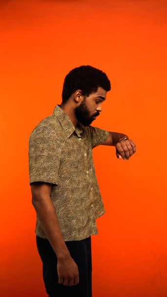 vertical view of young african american man with beard checking time on wristwatch on orange - Foto, immagini