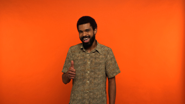 asombrado joven afroamericano con barba mostrando el pulgar hacia arriba sobre fondo naranja - Foto, Imagen