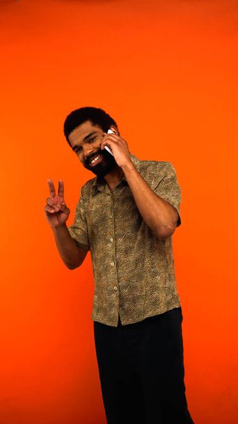 髭を生やしたアフリカ系アメリカ人男性がスマートフォンで語りオレンジで平和サインを表示します - 写真・画像