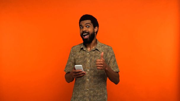 hombre afroamericano emocionado utilizando el teléfono inteligente y mostrando como en fondo naranja  - Foto, Imagen