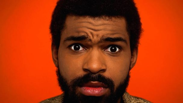 close-up van geschokte en bebaarde Afro-Amerikaanse man op zoek naar camera geïsoleerd op oranje  - Foto, afbeelding