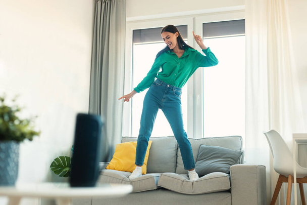 Happy Female Listening To Song Via Music Speaker Having Fun Dancing Standing On Sofa At Home On Weekend. Musical Device Advertisement Concept. Selective Focus - Фото, изображение