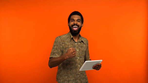 podekscytowany afrykański młody człowiek z brodą trzymający cyfrową tabletkę na pomarańczowym tle  - Zdjęcie, obraz