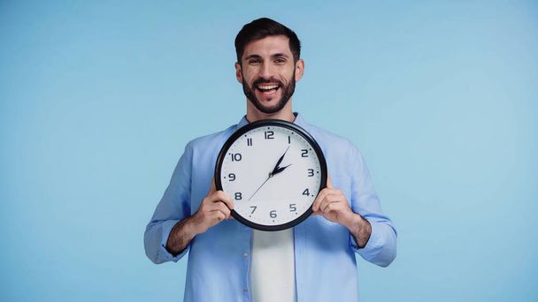 fröhlicher Mann im Hemd, Uhr in der Hand, während er isoliert in die Kamera auf blauem Hintergrund blickt  - Foto, Bild