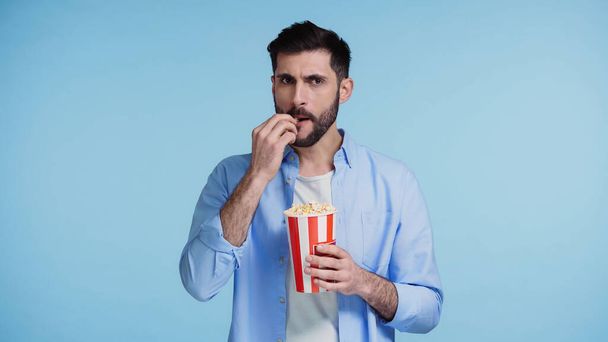 szakállas férfi kezében piros vödör és eszik popcorn elszigetelt kék - Fotó, kép