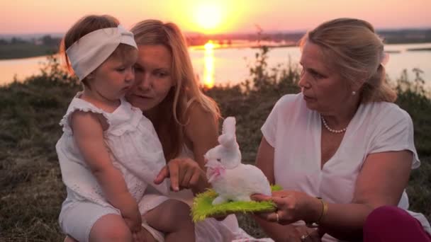 Family of only female members having a picnic on the hill at sunset - grandmother showing a bunny toy to little baby girl. Mid shot - Footage, Video