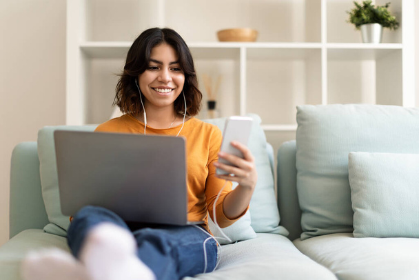 自宅でソファに座っている間にラップトップコンピュータとスマートフォンを使用して若いアラブ人女性を笑顔,携帯電話で友人と幸せな中東女性メッセージや新しいアプリを閲覧,スペースをコピー - 写真・画像