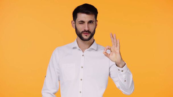 бородатый мужчина в белой рубашке показывает ОК знак в то время как подмигивание изолированы на желтый - Фото, изображение