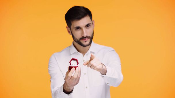 bearded man in white shirt holding box with engagement ring while pointing with finger at camera isolated on yellow - Foto, immagini