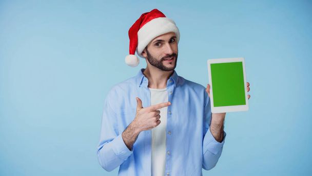 hombre feliz en sombrero de santa rojo apuntando con el dedo a la tableta digital con pantalla verde aislado en azul  - Foto, Imagen