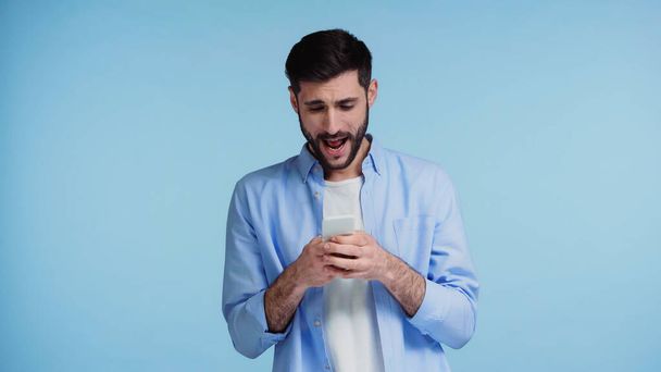 szakállas férfi használ okostelefon és sikoltozás közben üzenetküldés elszigetelt kék  - Fotó, kép