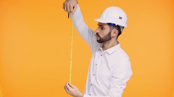soustředěný muž v bezpečnostní přilbě při pohledu na měřicí pásku izolovanou na žluté - Fotografie, Obrázek