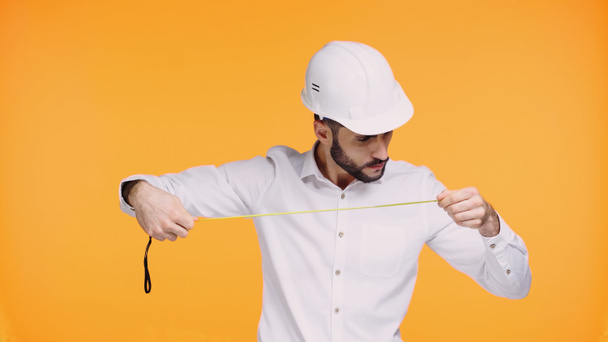 soustředěný architekt v klobouku při pohledu na měřicí pásku izolovanou na žluté - Fotografie, Obrázek