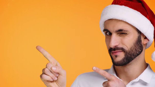 bearded man in red santa hat pointing away with fingers while winking isolated on yellow - Foto, Imagen