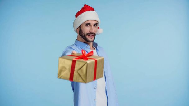 bearded man in red santa hat giving wrapped christmas present isolated on blue - Photo, Image
