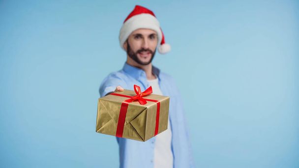 happy man in red santa hat giving wrapped christmas present isolated on blue - Fotografie, Obrázek