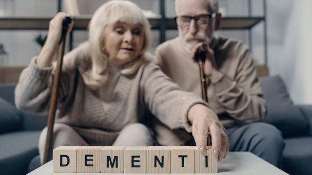 Senior man sitting near woman putting wooden cube with lettering and creating word dementia  - Photo, Image