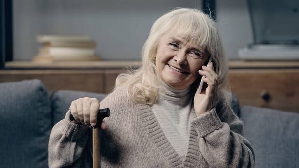 Улыбающаяся пожилая женщина с тростью, говорящая на смартфоне  - Фото, изображение