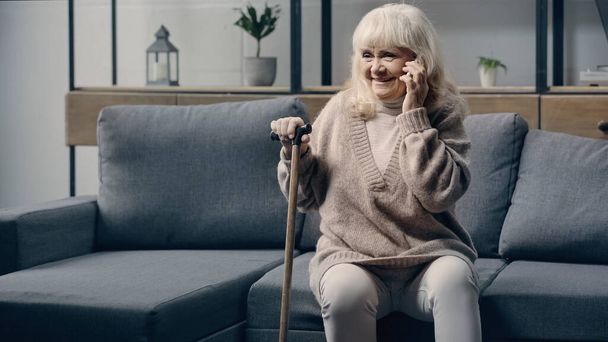 veselá starší žena s vycházkovou holí mluví na smartphone v obývacím pokoji  - Fotografie, Obrázek