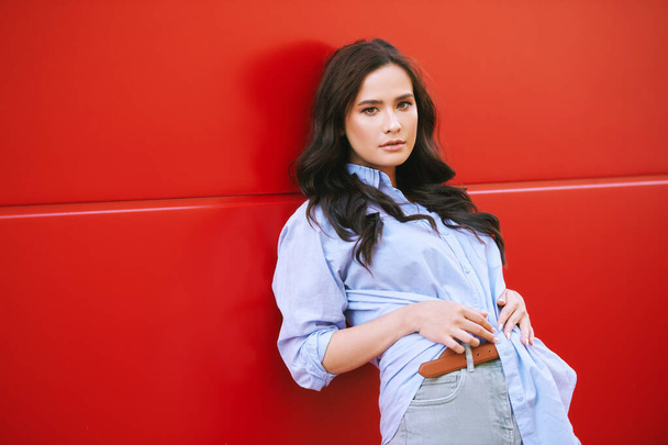 Ritratto di moda all'aperto di bella giovane donna appoggiata alla parete rossa, indossando camicia blu, trucco naturale e acconciatura ondulata - Foto, immagini