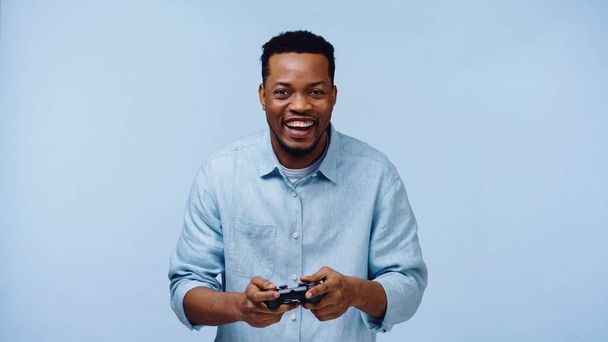 KYIV, UKRAINE - NOVEMBER 22, 2019: cheerful african american man playing video game isolated on blue  - Фото, изображение