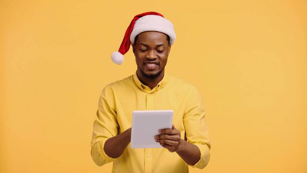 happy african american man in santa hat using digital tablet isolated on yellow - 写真・画像