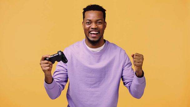 KYIV, UKRAINE - NOVEMBER 22, 2019: positive african american man holding joystick and rejoicing isolated on yellow  - Foto, afbeelding