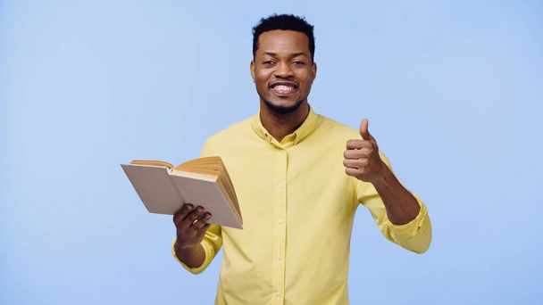 счастливый африканский американец в желтой рубашке показывая большой палец вверх и держа книгу изолированы на синий - Фото, изображение
