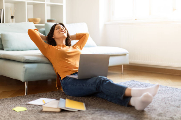 Portrait einer glücklichen jungen Araberin, die sich nach dem Online-Studium mit Laptop und Arbeitsbüchern auf dem Boden entspannt, eine fröhliche Frau aus dem Nahen Osten, die mit den Händen hinter dem Kopf ruht, Distanzlernen genießt und Raum kopiert - Foto, Bild