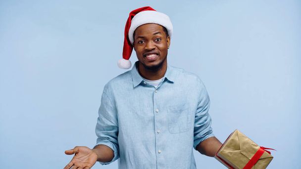 verwirrter afrikanisch-amerikanischer Mann mit Weihnachtsmütze, Weihnachtsgeschenkschachtel und achselzuckender Geste auf blauem Grund - Foto, Bild