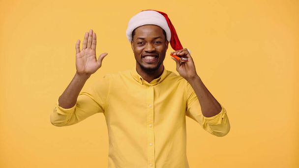 szczęśliwy Afrykanin amerykański człowiek w Santa kapelusz macha ręką odizolowany na żółty  - Zdjęcie, obraz