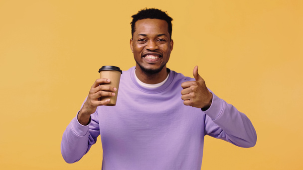 cheerful african american man in purple pullover holding coffee to go and showing like isolated on yellow - Zdjęcie, obraz