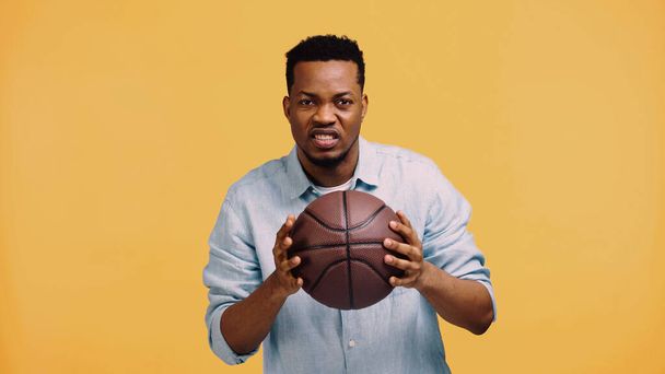 displeased african american man holding basketball isolated on yellow - Foto, imagen
