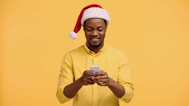黄色に隔離されたスマートフォンを使ってサンタ・ハットの幸せなアフリカ系アメリカ人男性 - 写真・画像