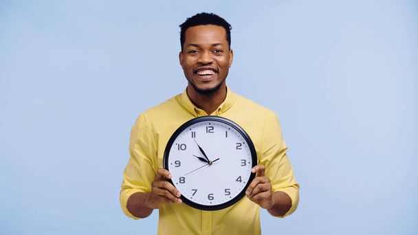 wesoły Afrykanin amerykański człowiek trzymając zegar i patrząc na aparat odizolowany na niebiesko - Zdjęcie, obraz