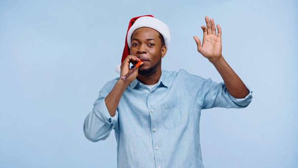 Afrikanisch-amerikanischer Mann mit Weihnachtsmannhut bläst Partyhorn und gestikuliert isoliert auf blau - Foto, Bild